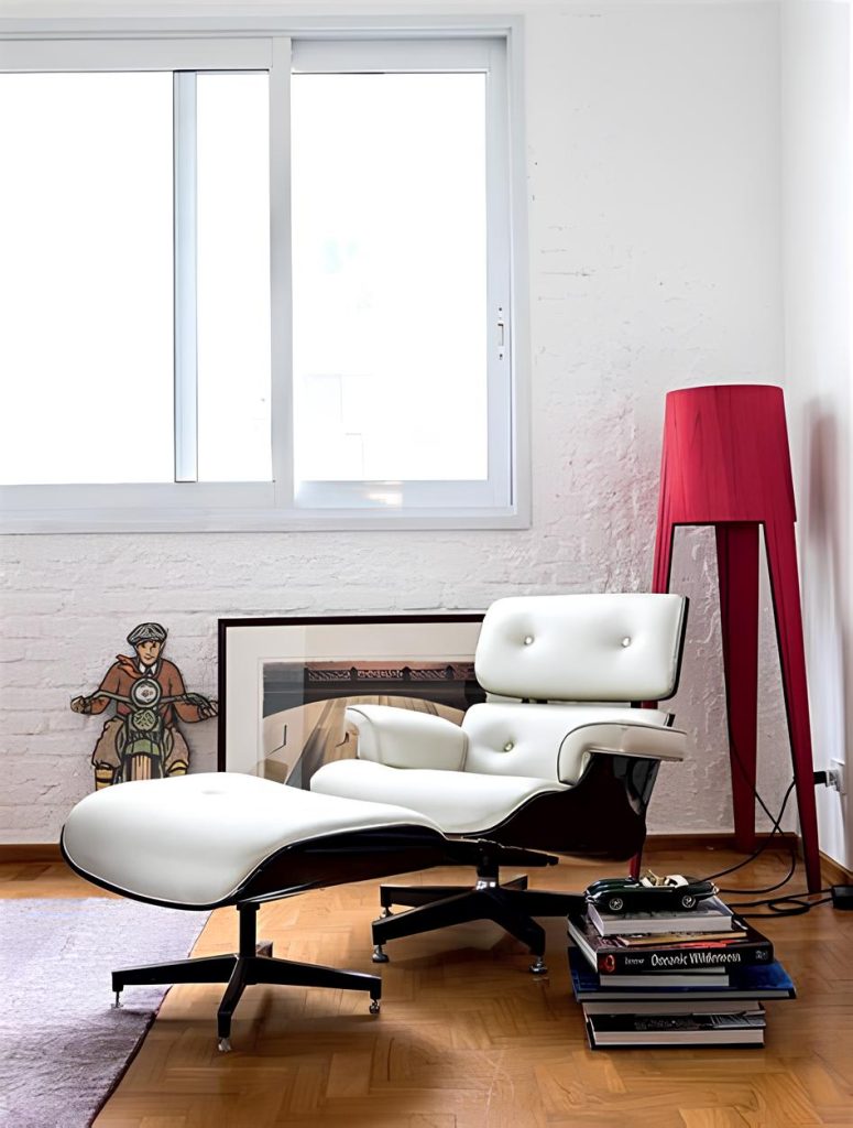 ambiente com uma poltrona branca modelo charles eames de couro estilo giratório, atrás uma luminária vermelha moderna ao lado quadro e uma janela com luz natural.
