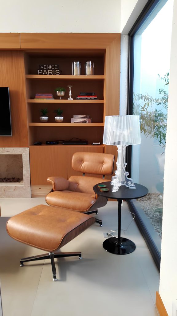ambiente com uma poltrona de couro marrom modelo cherles eames, ao lado uma mesinha de metal preta com uma luminária transparente, atrás uma estante de madeira moderna marrom com vários objetos decorando.