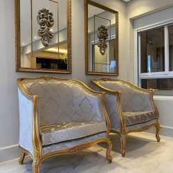 dois modelos de poltronas estilos Luiz XV de cor bege claro com alto relevo e detalhes de metal dourado, atrás dois quadros espelhados com uma moldura central 3D.