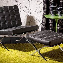 poltrona com modelo barcelona preta com um descanso de pés acompanhando ao lado uma mesinha redonda com pés altos na cor verde com vasos decorativos em cima dela.