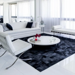 sala de estar ampla com três poltronas, sendo duas estilos cadeiras com pés de metal e estofamento branco e a outra poltrona com três lugares na cor branca no chão um tapete grande escuro, em cima uma mesinha de centro redonda baixa branca com objetos em cima para decorar, ao lado das poltronas uma sofá grande com almofadas decorativas.