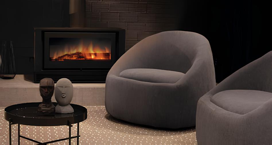 sala de estar com 2 poltronas magnólias nas cores cinzas, ao fundo uma lareira elétrica e na frente uma mesinha de centro redonda com objetos decorativos 