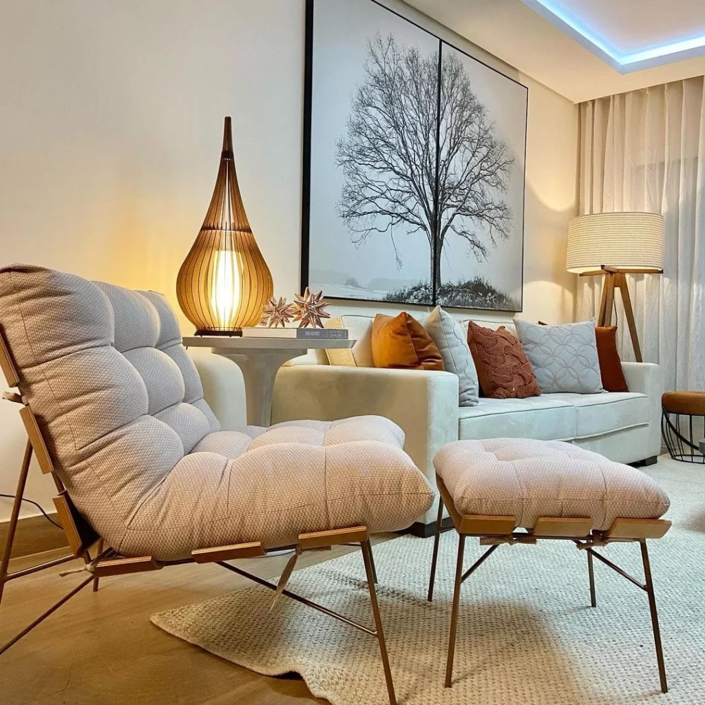 sala de estar com uma poltrona magnólia clara, ao lado sofa claro de 3 lugares com almofadas decorativas e no meio dos dois uma mesinha redonda com luminárias e outros objetos decorativos 