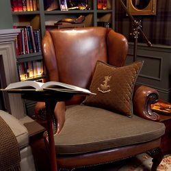 sala de leitura com uma poltrona antiga de couro na cor marrom, ao lado um pedestal pequeno com um livro, atrás uma prateleira grande com vários livros.