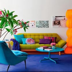 sala de estar bem colorida com um sofa encostado na pare na cor amarelo esverdeado com algumas almofadas coloridas em cima , na frente uma mesinha de centro redonda na cor azul com objetos em cima decorando e uma poltrona azul com estofado na mesmo cor, do lado esquerdo plantas para decorar ambiente.