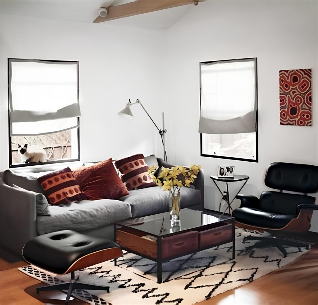 sala de estar com poltrona charles eames preta de couro, sofa tres lugares na cor cinza com almofadas decorativas, no centro uma mesa com gavetas e vidro em cima dela um vaso de vidro com flores amarelas, e um tapete branco com desenho preto