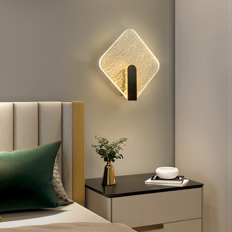 quarto com cama de casal, ao lado um criado mudo com um vasinho de planta e livros em cima dela, na parede atrás da cama uma luminária em formato de abajur moderna.