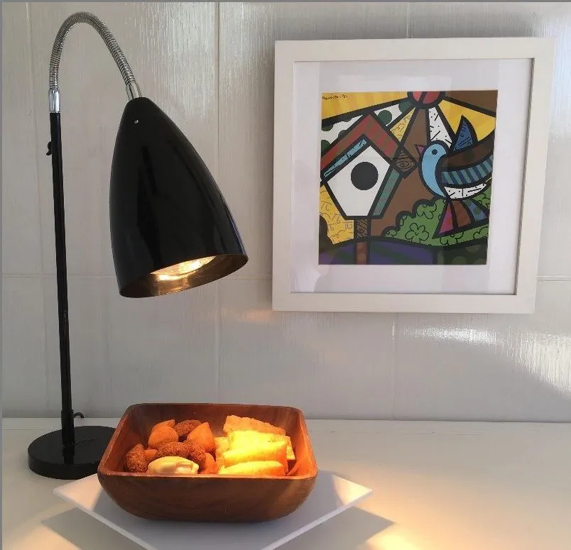 mesinha com uma luminária reclinável na cor preta, em baixo dela um potinho de madeira com comidas dentro dele, atrás um quadro decorativo na parede.