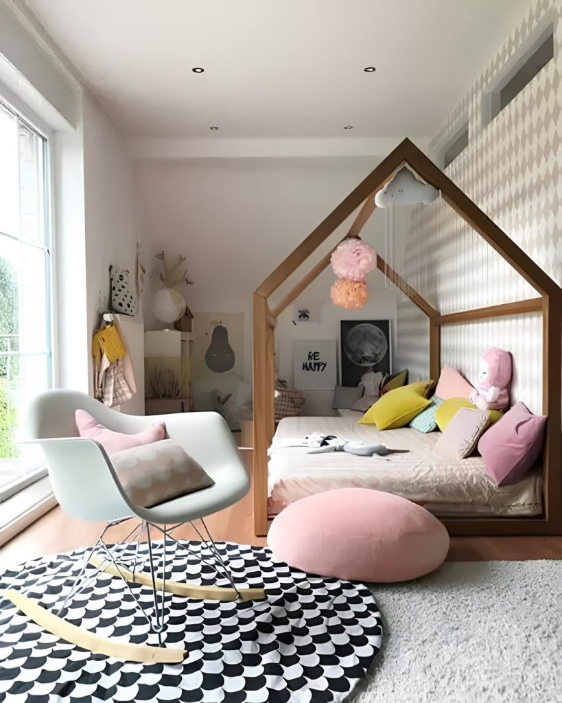 quarto infantil com cama feita em formato de casinha em madeira com travesseiros decorando, ao lado uma cadeira de balanço com almofadas em cima, ao chão um tapete redondo nas cores preto e branco, ao fundo quadros na parede e prateleiras com objetos.