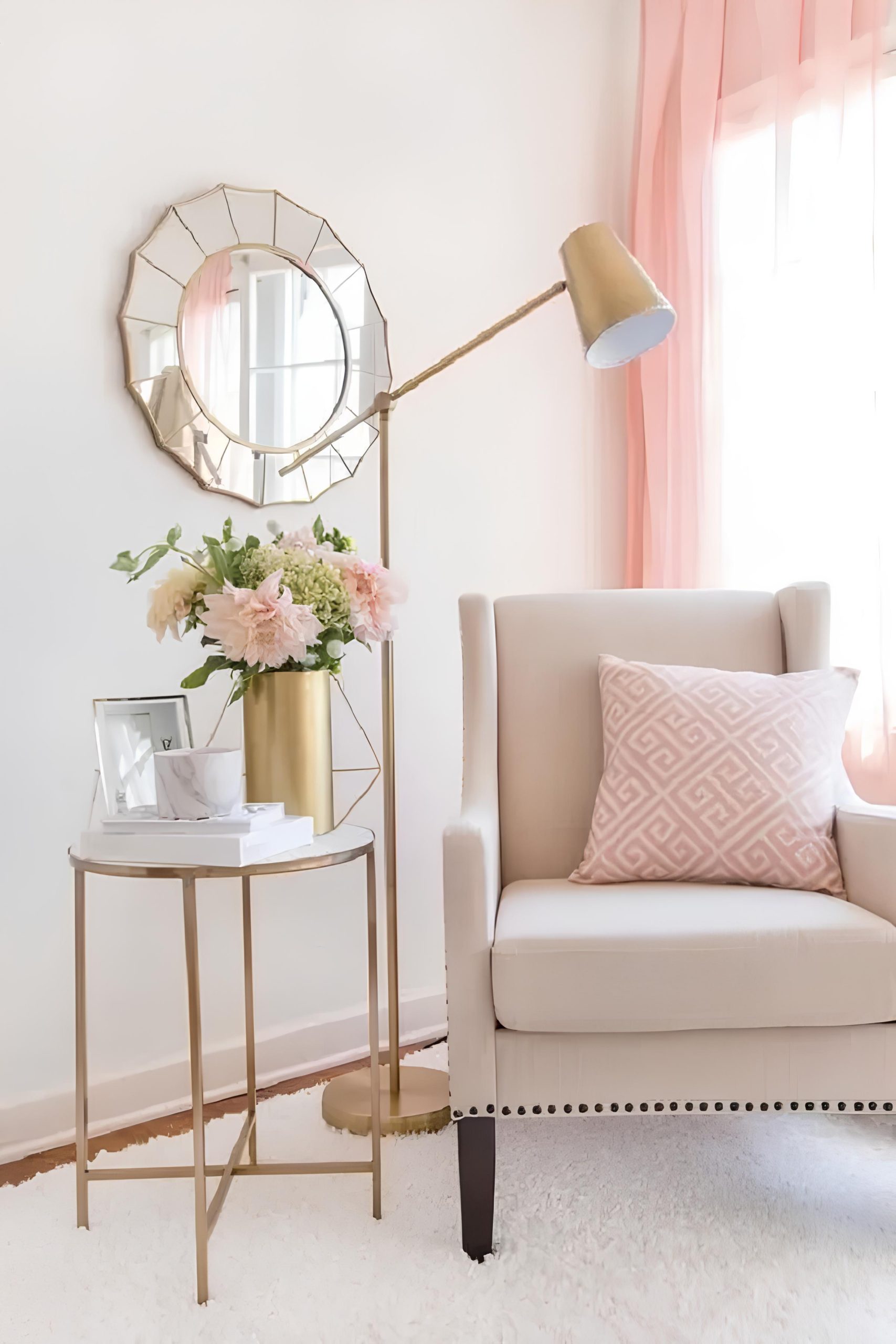 espaço com um sofá claro, com almofada em cima, ao lado dele uma mesinha redonda com vaso de rosas e outros objetos decorando, atrás uma luminária de chão, na parede atrás do sofá um espelho pendurado na parede oval .