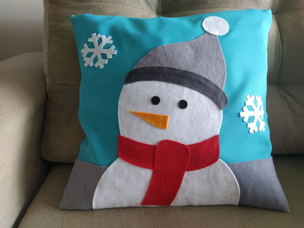 modelo de uma almofadas confeccionada de feltro com um boneco de neve e tema natalino em cima de um sofá na cor bege.