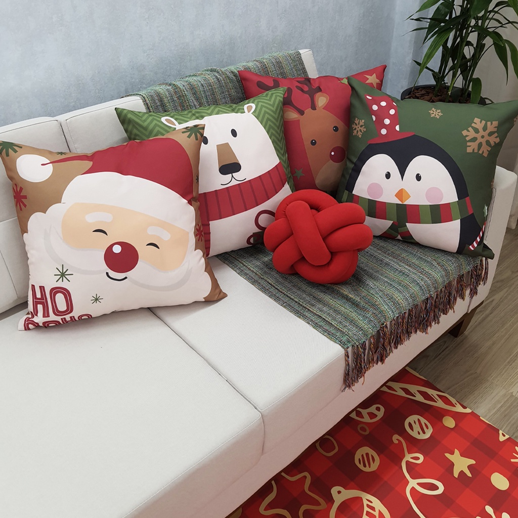 sofa na cor branca com pés de madeira, em cima uma manta esverdeada com quatro almofadas temáticas de natal, ao lado um vaso grande com planta.
