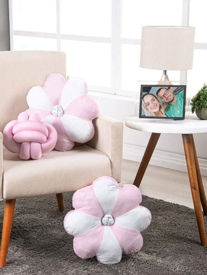 ambiente com uma poltrona rosa de pé palito com almofadas diferentes infantis em cima, ao lado uma mesinha redonda de madeira com porta retrato e vasinho de planta, no chão na frente da poltrona uma almofada com formato de flor nas cores rosa e branco.