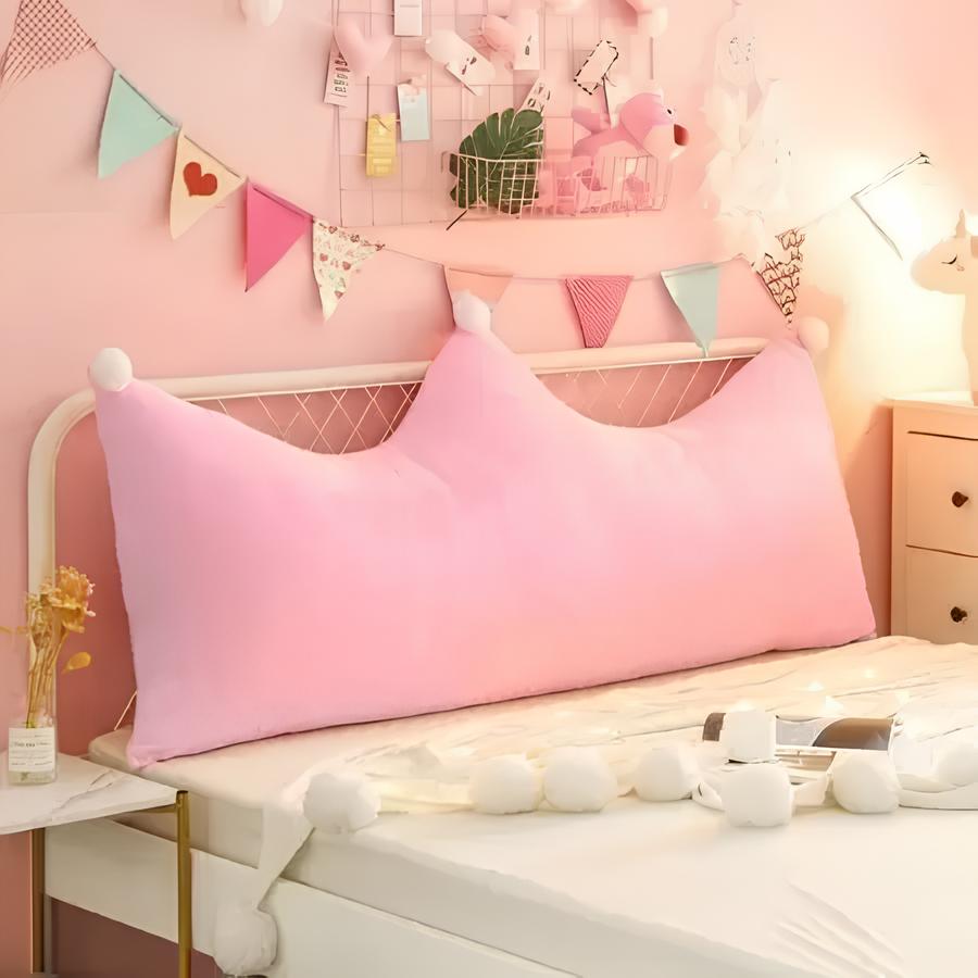 quarto infantil de menina com cama de solteiro branca e uma almofada grande em formato de coroa de rainha na cor rosa, nas laterais um criado mudo com gavetas branco e uma luminária de unicórnio e do outro lado uma mesinha de mármore quadrada com vasinho de planta para decorar.