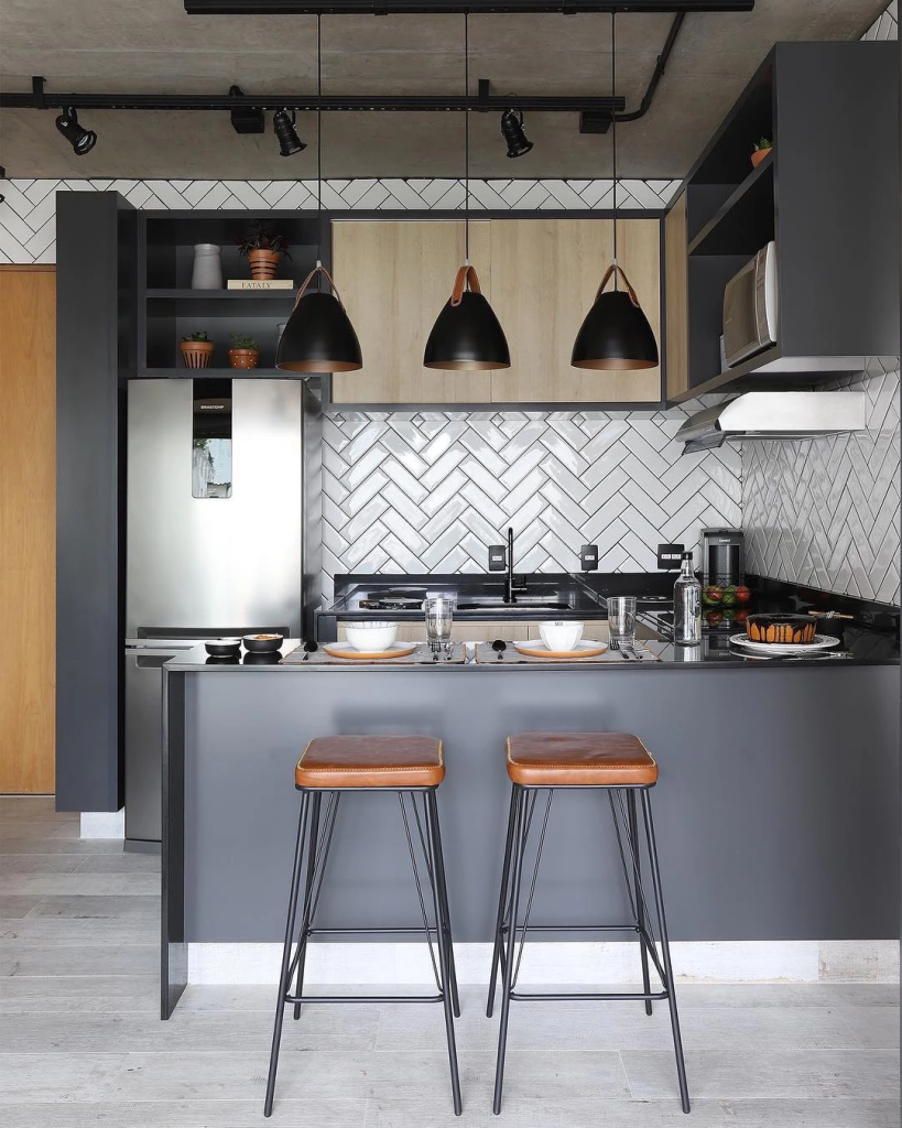 cozinha pequena com móveis planejados nas cores marrom, preto e cinza, com bancada na mesma cor, xícaras e copos em cima dela, banquetas em frente a bancada, em cima no teto três luminária pendentes na cor preta.