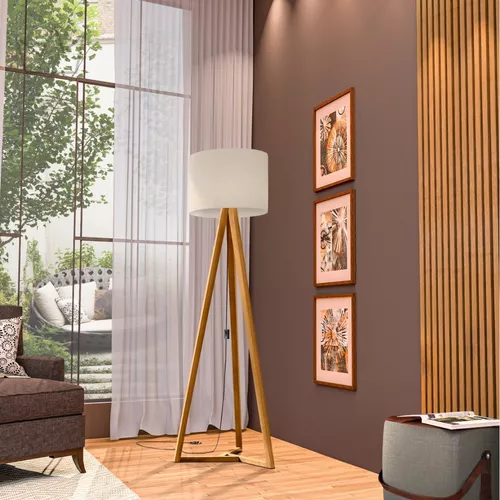 sala com uma luminária gigante de pés de madeira, na parede ao lado dela três quadros na parede decorativos, ao lado uma poltrona marrom com almofadas em cima decorando, atrás uma janela grande de vidro com vista de uma poltrona de descanso de varanda.
