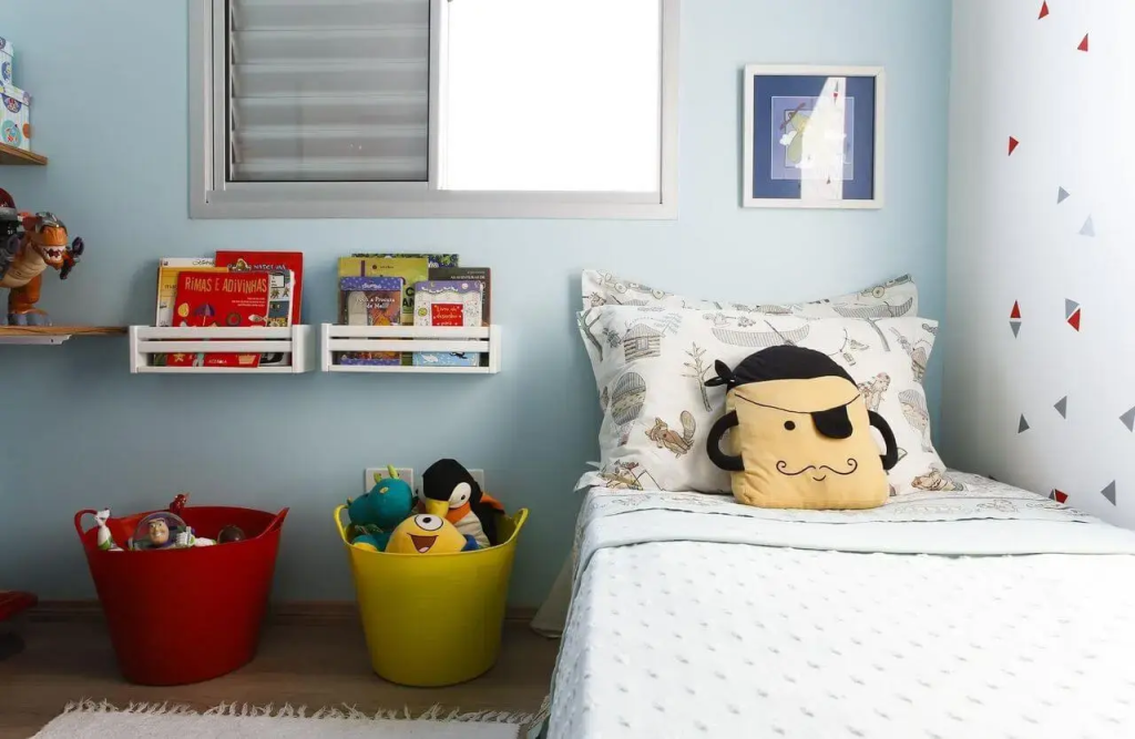 quarto infantil de menino com cama de solteiro, em cima dela uma almofada em formato de pirata ,ao lado da cama dois baldes em cores amarelo e vermelho com vários bichinhos de pelúcia dentro. em cima duas prateleiras com livros dentro.