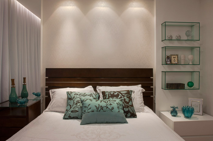 quarto com cama de casal, com um criado mudo ao lado dela e objetos em cima dele decorando, na parede nichos de vidro e no teto luminária embutida.