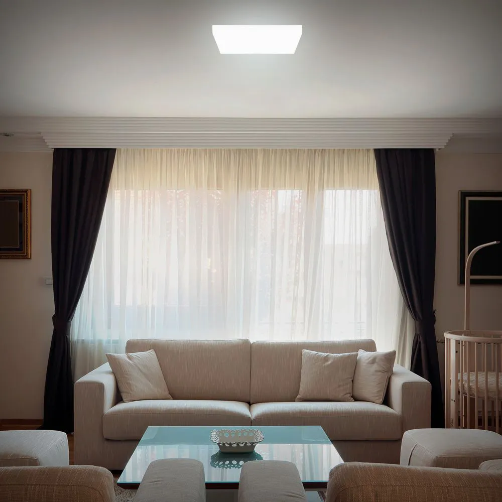 sala de estar com sofá e poltronas nas mesas cores, ao meio deles uma mesinha de centro quadrada de madeira, atrás do sofa janela com cortinas brancas e marrom escuro, no teto uma luminária embutida.