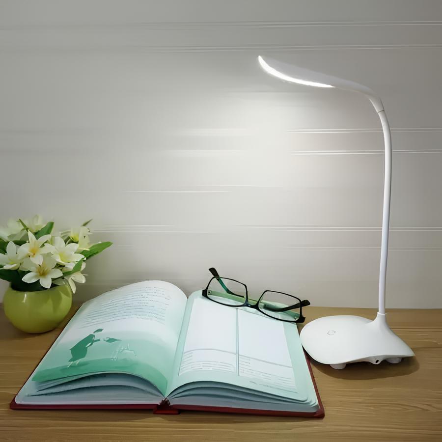 mesinha de madeira com um livro e óculos de leitura em cima dela, de uma lado um vasinho decorativo com flores e do outro uma luminária de LED.