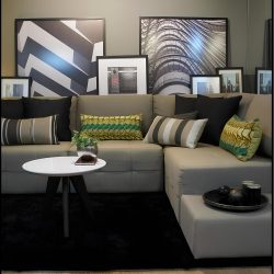 sala de estar ampla e moderna com vários quadros na parede de tamanhos grandes, médios e peqeunos, em frente aos quadros um sofá na cor cinza em estilo L com várias almofadas em cima decorando, na frente do sofá uma mesinha redonda branca com pés na cor preta, em cima da mesinha vasinho com plantas e alguns objetos decorativos.