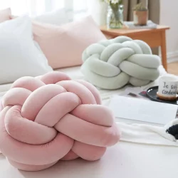 cama de casal com almofadas em cima decorando das cores rosa claro e branco, duas delas em formatos de almofadas de nó, ao lado da cama uma mesinha de maderia quadrada com objetos decorativos em cima dela.