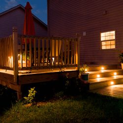 área externa com acessoa para um deck de madeira com mesas e cadeiras, nas escadas luminárias de led, ao redor gramado e vasos com flores.