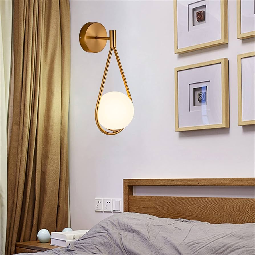 quarto com cama de casal, e cabeceira de madeira, ao lado da cama um criado mudo com livros em cima dele, na parede atrás da cabeceira quadros decorativos e uma luminária de parede estilo LEd moderna.