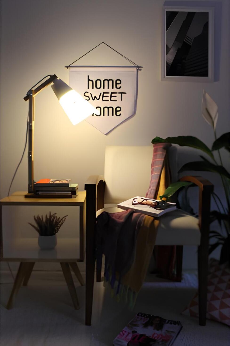 ambiente com uma cadeira com manta, livro e um óculos em cima dela, ao lado uma mesinha quadrada de madeira com livros e uma luminária em cima dela, atrás da cadeira uma parede com quadro pendurado, e do outro lado um vaso com planta da espécie copo de leite.