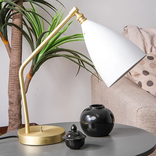 mesinha redonda na cor escura com uma luminária de mesa dourada com branca e dois objetos decorando, ao fundo um sofá na cor creme com almofadas, atrás da mesinha uma planta alta.