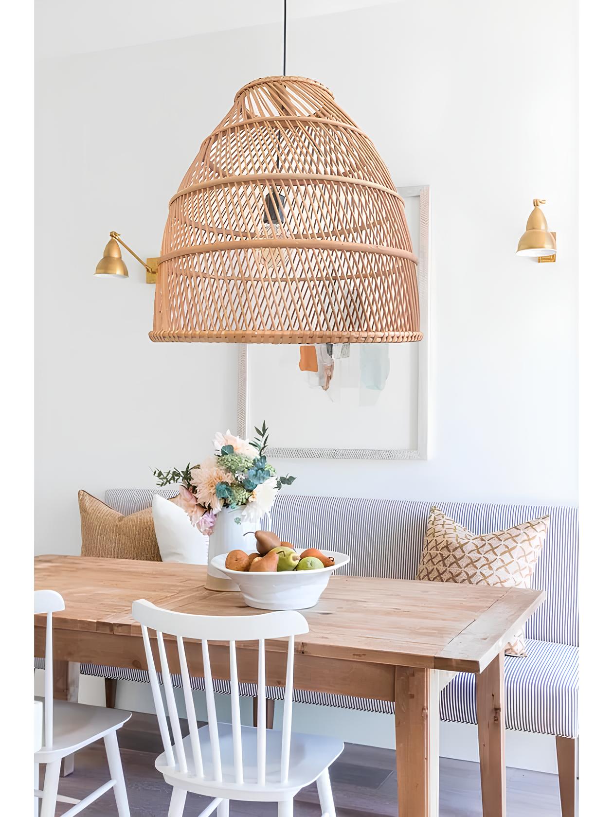 ambiente com um sofá com listras finas zuis e branca, almofadas cremes e brancas em cima dele, na frente do sofá uma mesa de madeira com duas cadeiras brancas de madeira e em cima da mesa um vaso com flores e travessa com frutas em cima no centro da mesa no teto um lustre pendente de vime.