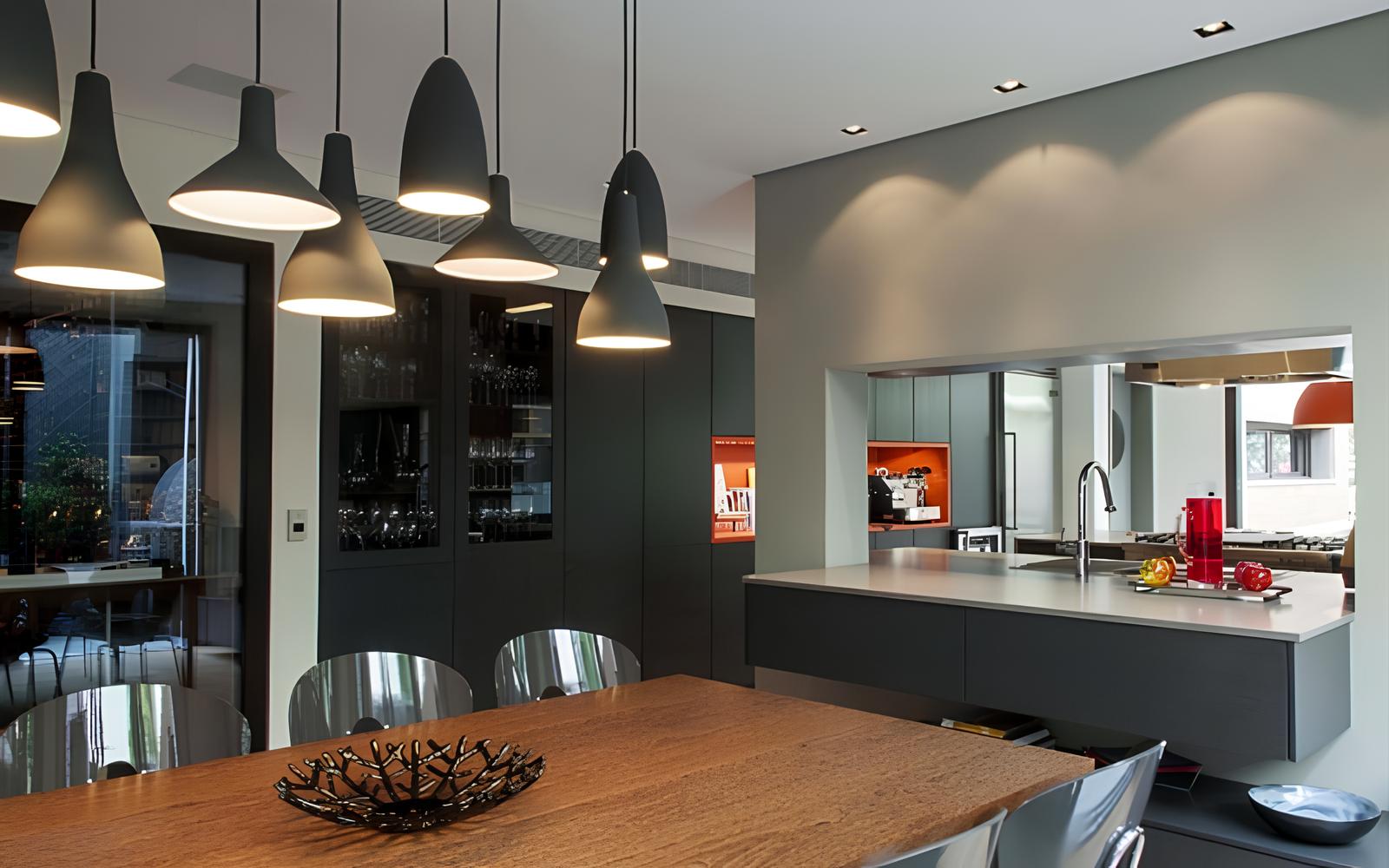 cozinha moderna em tons cinza escuro e preto, com mesa de madeira e cadeiras de inóx, na lateral uma armário planejado na cor preta com vidros, e uma pia também planejada, luminárias pendentes no centro da mesa no teto.