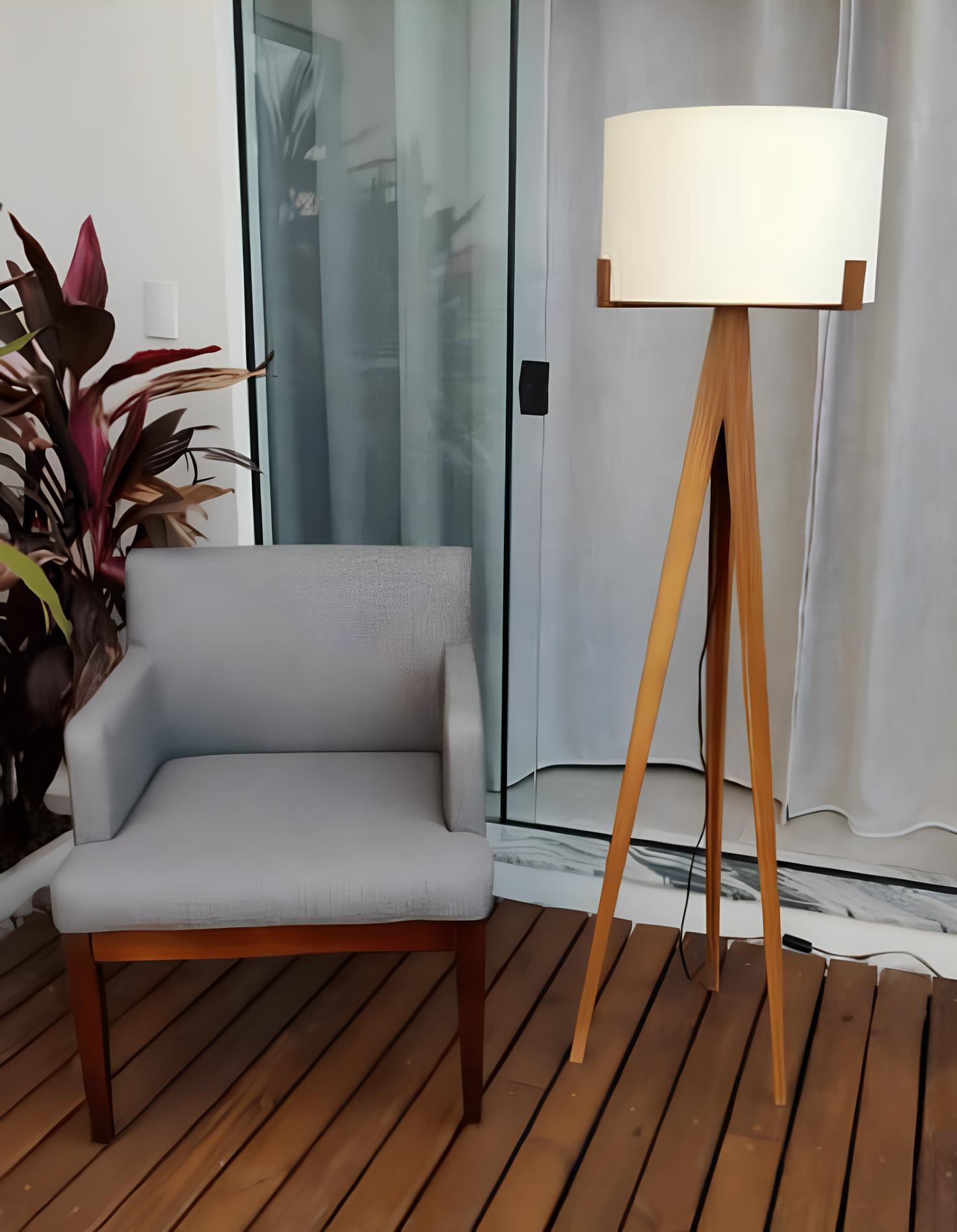 ambiente com uma poltrona na cor cinza, ao lado uma luminária de chão alta, ao lado da poltrona um vaso com planta grande, atrás da poltrona uma porta de vidro grande com cortinas brancas.