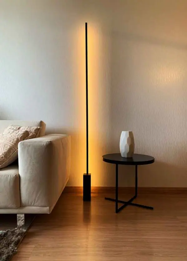 ambiente com uma sofá e almofadas em cima nas cores claras, ao lado uma luminária LED de chão e ao lado uma mesinha redonda com vasinho decorativo.