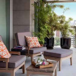 varanda com duas poltronas de madeira com estofamento marrom com almofadas em cima na frente delas uma mesinha da mesma cor e estilo das poltronas, ao lado dois vasos grandes redondos com plantas para decorar.