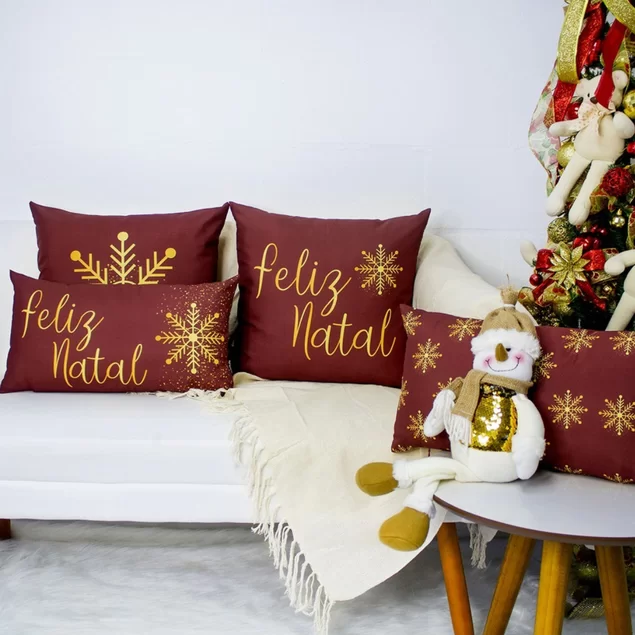 ambiente com sofá branco com manta branca em cima e almofadas natalinas, ao lado uma mesinha redonda de madeira branca com uma almofada natalina e um boneco de neve de pano natalino, atrás uma arvore de natal com enfeites.