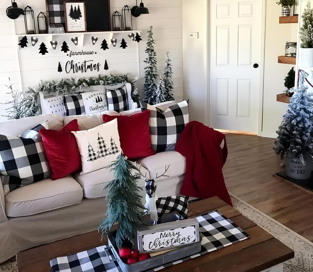 sala de estar com um sofá claro com almofadas decorativas de natal, atrás dele um banco na cor branca com almofadas brancas e xadrez nas cores branco e preto, em cima uma prateleira com objetos decorativos, ao lado ao sofá uma árvore natalina e em frente ao sofá uma mesinha de madeira com objetos natalinos decorando.