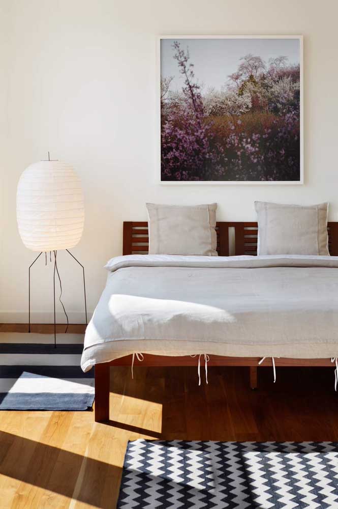 quarto com cama de casal de madeira com cabeceira de madeira, roupas de cama na cor clara, ao lado da cama uma luminária de chão alta, grande e moderna, na parede atrás da cama uma quadro decorativo grande na parede.
