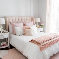 quarto com cama de casal, cabeceira na cor rosa, almofadas nos tons rosa e branco, nos pés da cama uma manta na cor rosa claro, nas laterais criados mudos redondos com gavetas de madeira na cor branco com luminárias e objetos decorativos em cima.