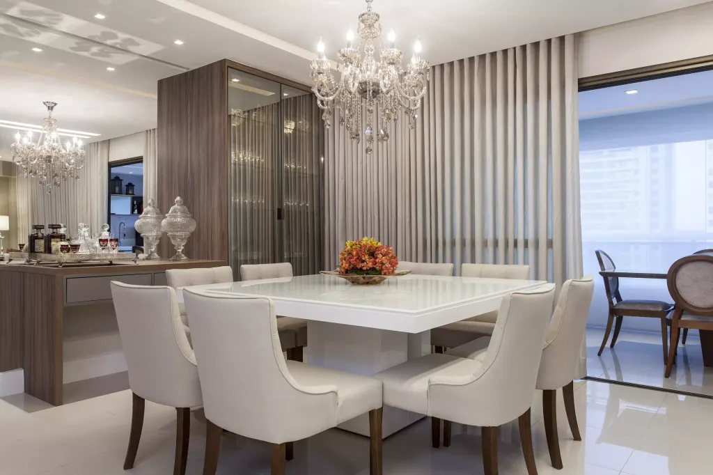 sala de jantar com mesa grande e cadeiras nas cores branca, com flores em cima dela, ao lado da mesa uma mesinha de madeira MDF com jarras e copos com de vidros, no centro ao teto um lustre de cristal, ao fundo da mesa uma cortina grande branca e atrás mesas e cadeiras na varanda fechada.