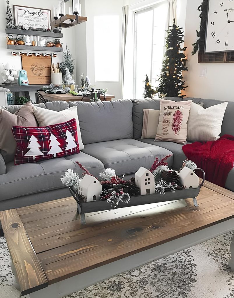 sala de estar com um sofá em L na cor cinza com almofadas de natal e uma manta vermelha em cima, em frente ao sofá uma mesinha quadrada de madeira com enfeites natalinos, atrás do sofá uma árvore com enfeites de natal, uma mesa de jantar enfeitada para natal.