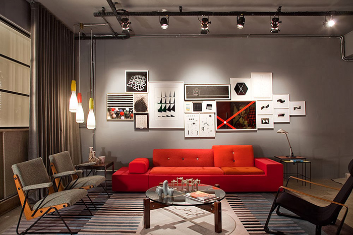sal de star moderna ampla com sofá vermelho, poltronas estilos cadeiras, no centro uma mesinha redonda de vidro com pés de madeira e objetos decorativos em cima, atras na parede ao fundo do sofa quadro decorativos na parede, nas laterais e em cima no teto luminárias penduradas.