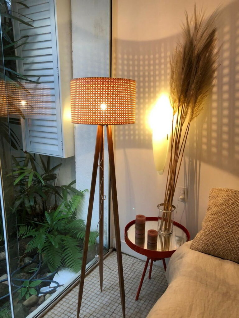 espaço com um sofá bege, ao lado uma mesinha redonda de vidro com objetos decorativos em cima incluindo pelo de pampa, ao lado uma luminária alta e trás dela uma porta de jardim de inverno de vidro com plantas a vistra.