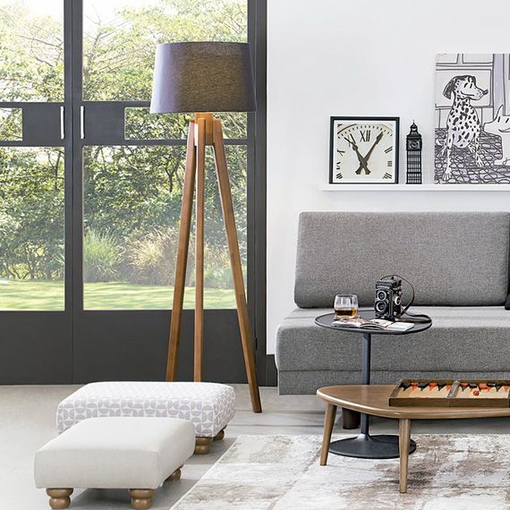 sala de estar com um sofá na cor cinza, ao lado dele uma luminária alta de pés de madeira, em frente um mesinha de madeira redonda, atrás uma porta de metal com vidro e vista para um jardim.